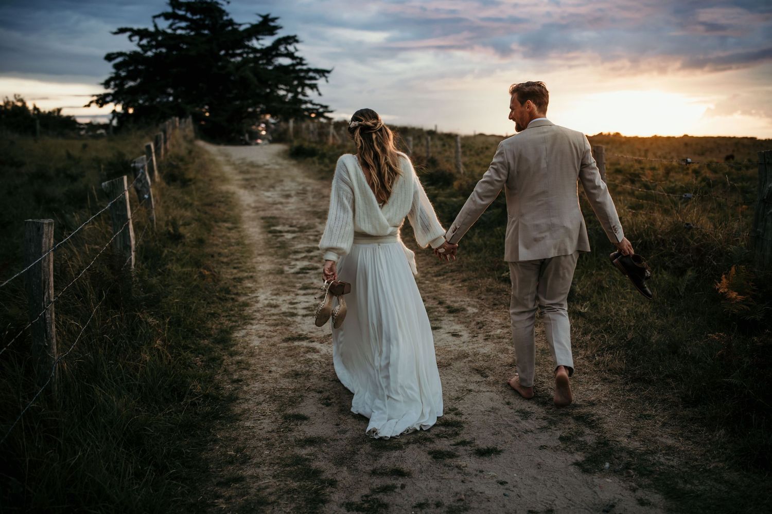 A couple walks hand in hand down a dirt path at sunset in their wedding attire.