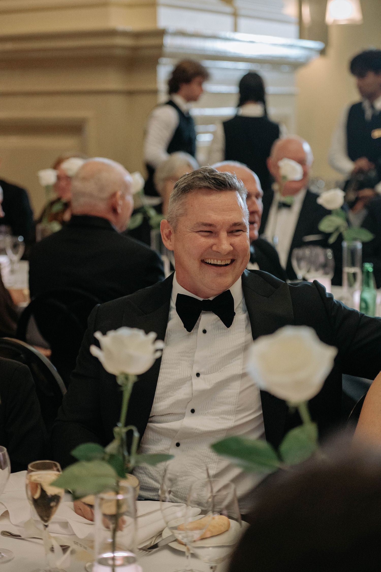 Someone in a black tuxedo and bow tie laughs joyfully at an elegant dinner reception with white roses on the table.