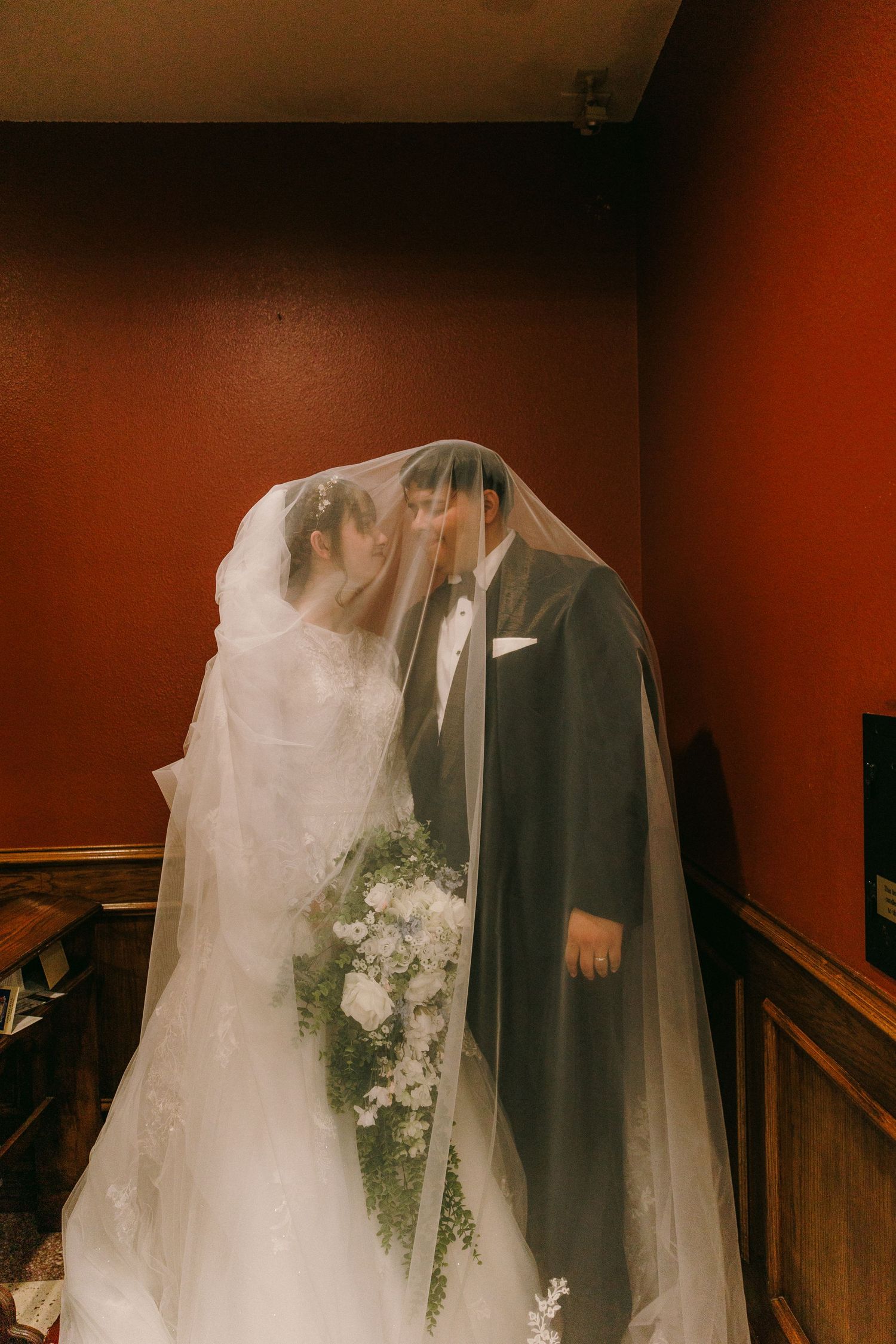 A couple on their wedding day wearing formal attire with a long white veil and holding a white floral bouquet.