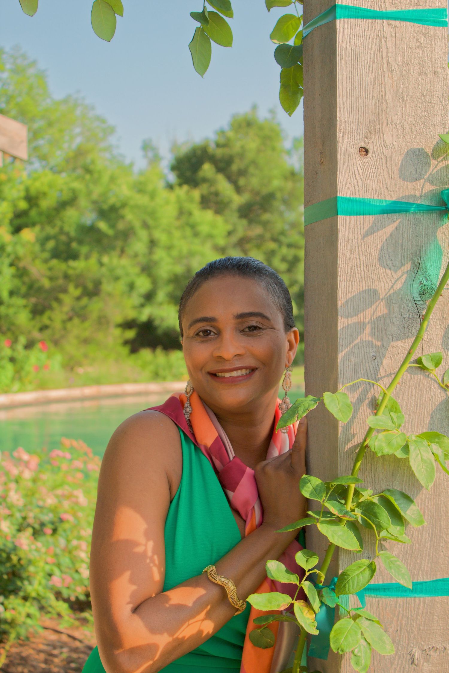 Smiling person in green tank top poses near a garden post with climbing vines on a sunny summer day.