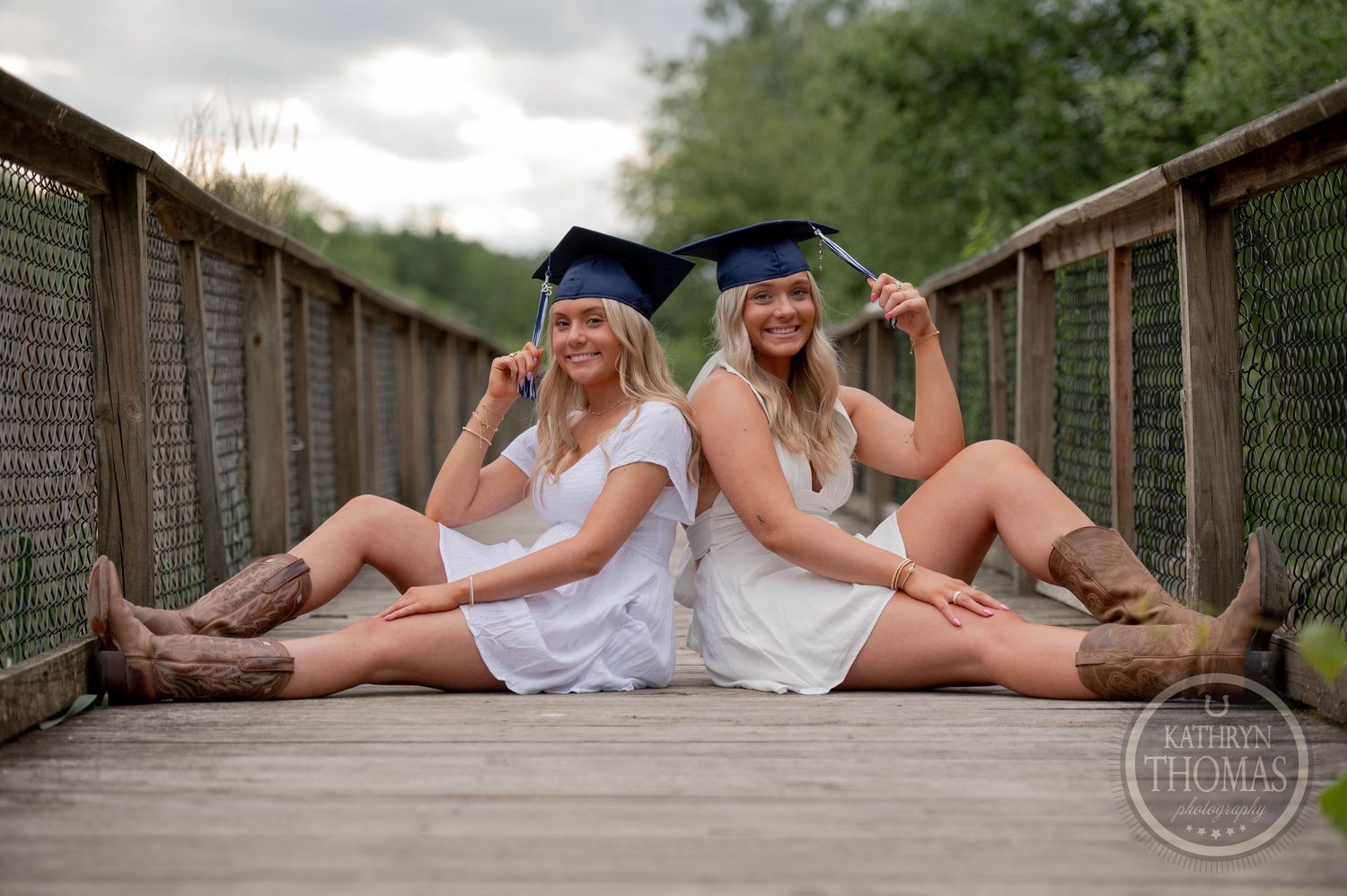 Two graduates in white dresses and graduation caps sit back-to-back on a wooden bridge walkway in nature.