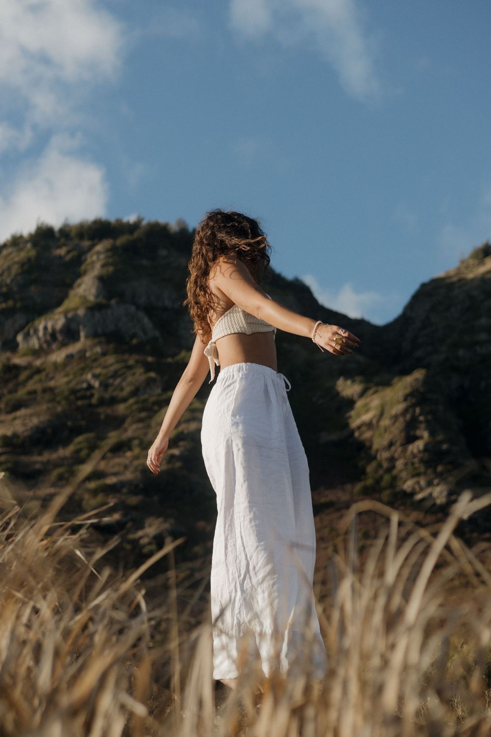 A person in white flowing clothing stands in tall grass against a mountainous backdrop on a sunny day.