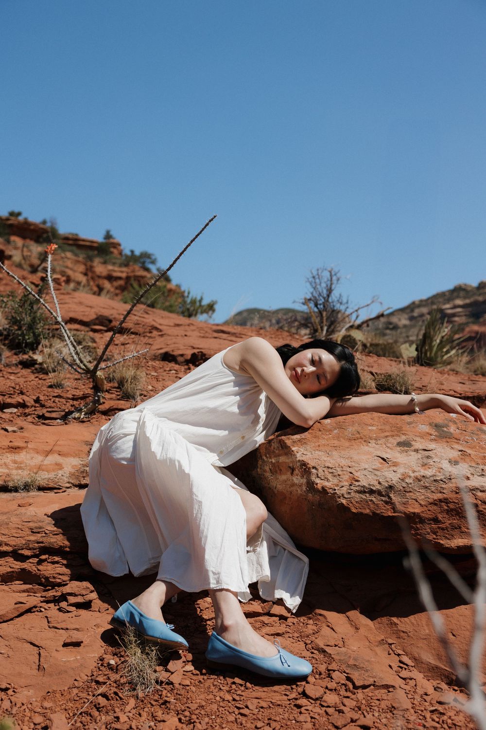 Someone in a flowing white dress and blue flats reclines on red rock formations in a desert landscape beneath blue sky.