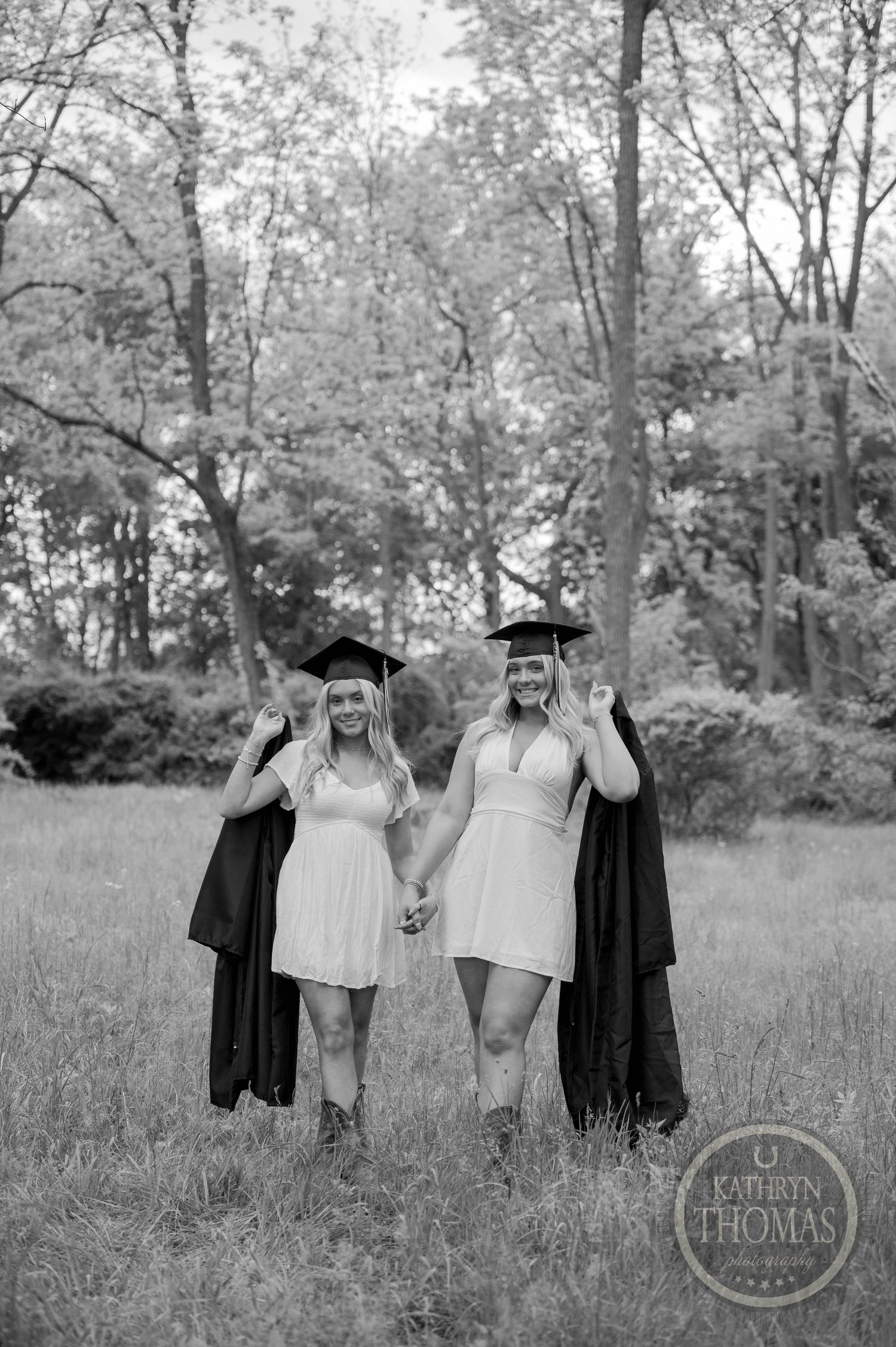 Two graduates in matching navy gowns celebrate in a lush green field surrounded by spring trees.