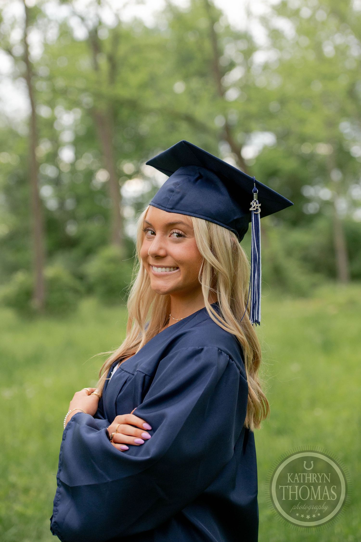 A graduate in navy blue cap and gown poses outdoors with a bright smile against a green natural background.