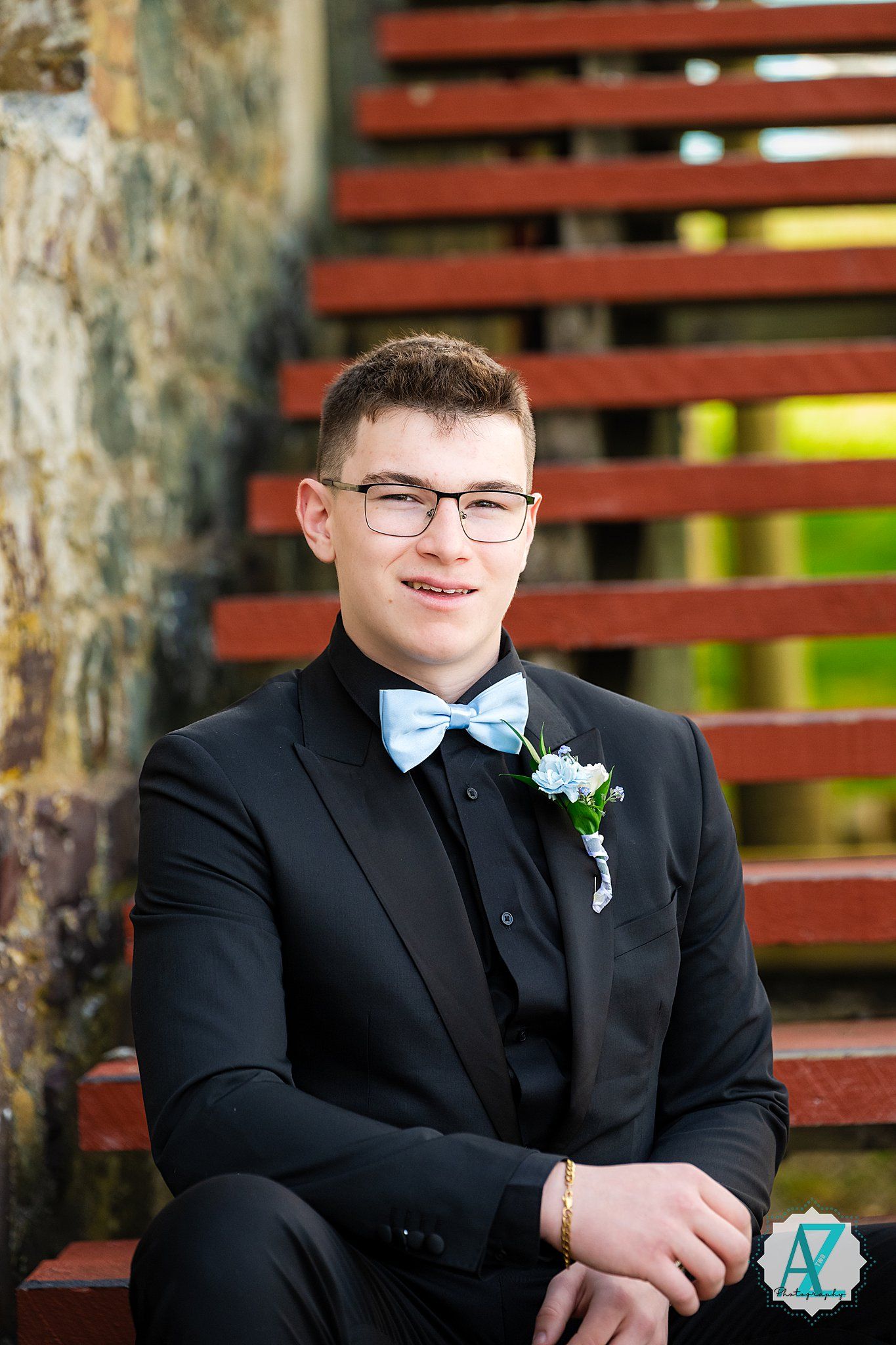 A young person in a formal black tuxedo with a light blue bow tie and boutonniere sits on red wooden steps.