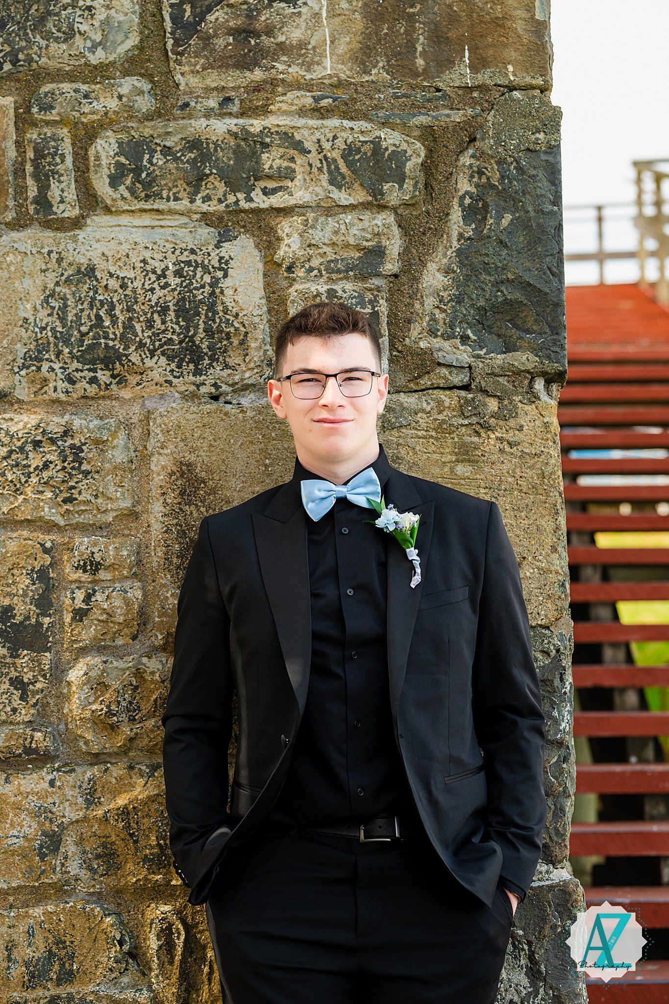 A formal portrait against a stone wall features a person in a black tuxedo with bow tie and boutonniere.