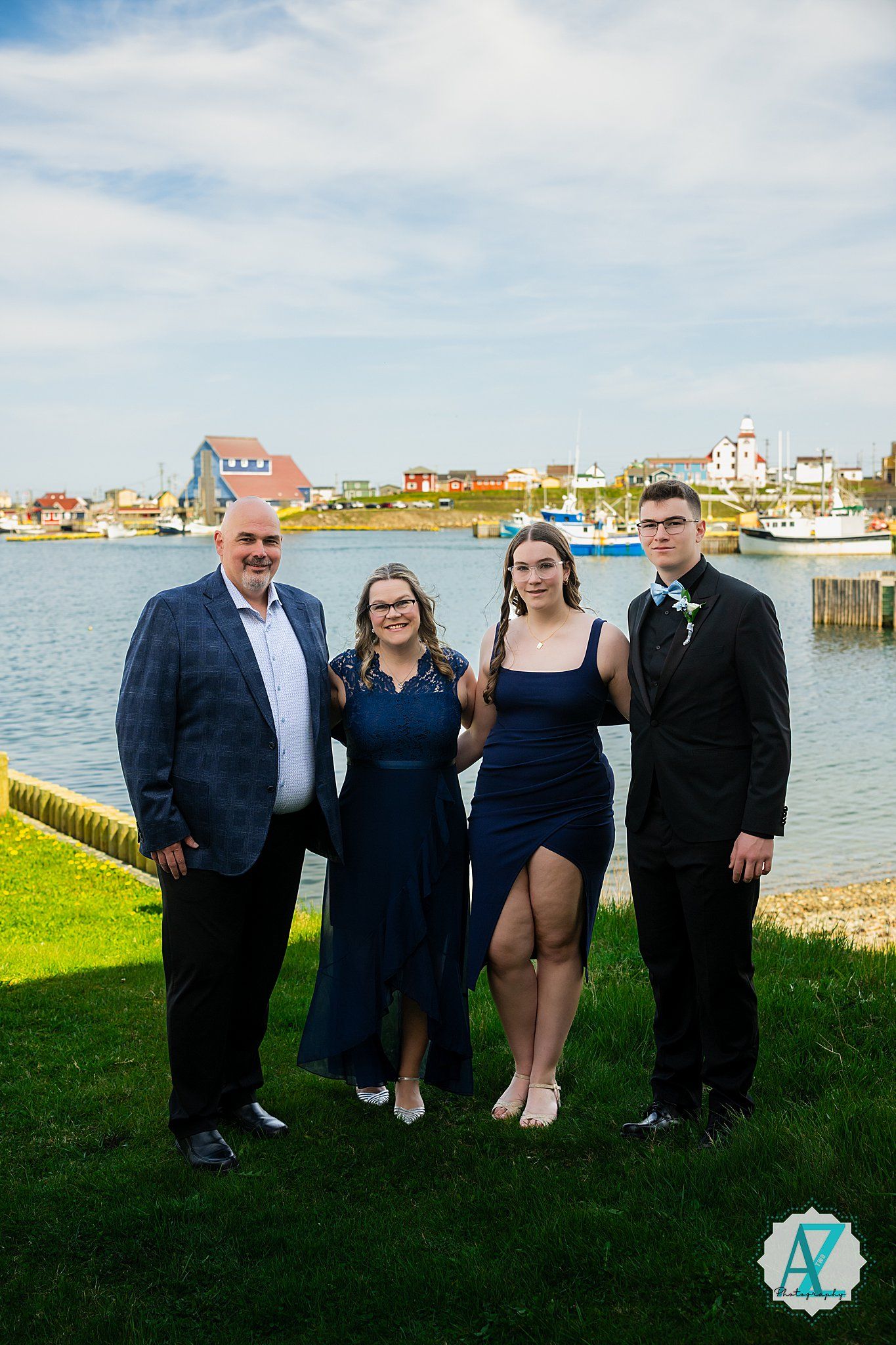 A group in formal attire stands together on grass near a harbor with coastal buildings and boats behind them.