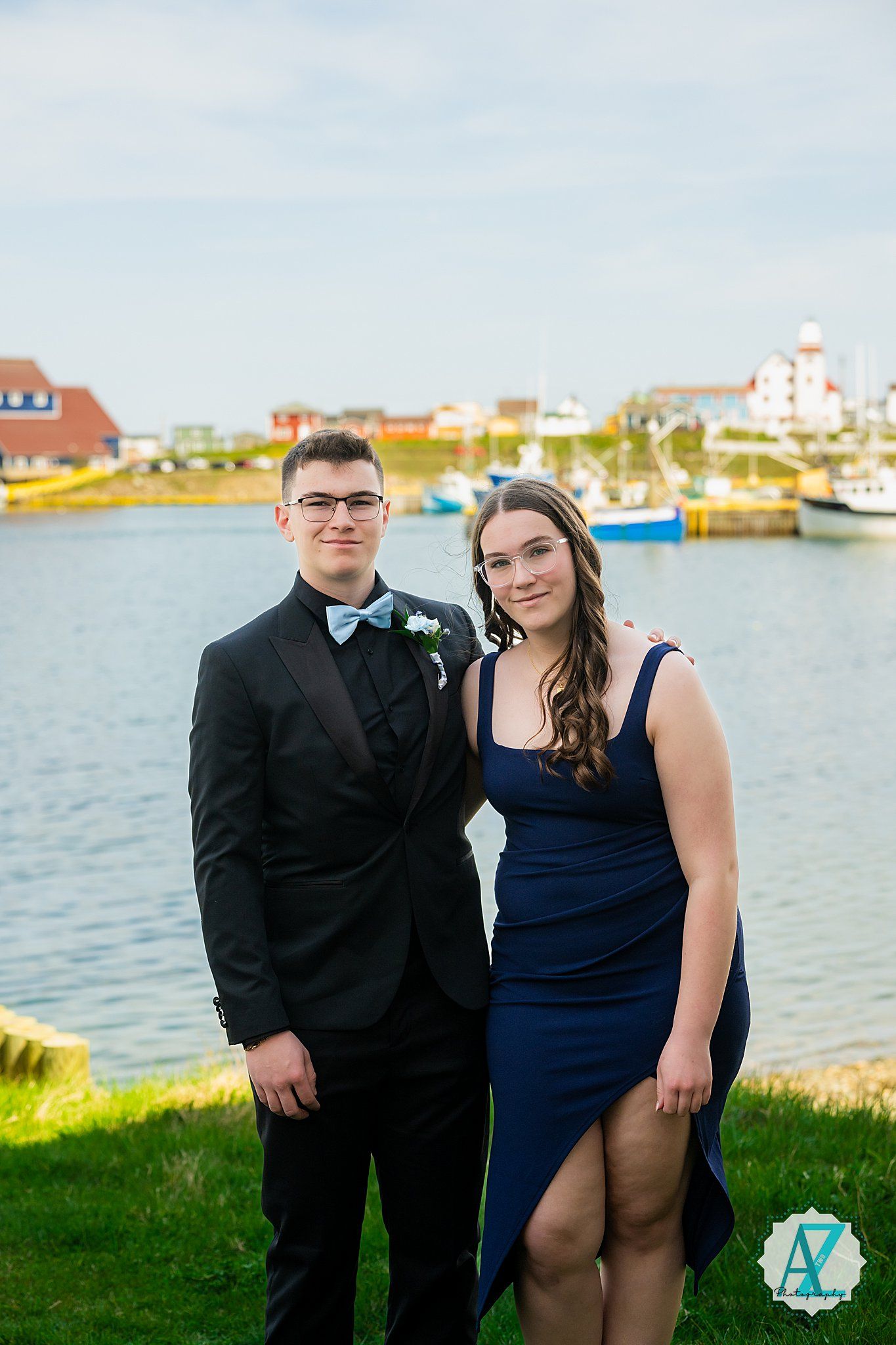 A formal couple poses for a photo by a scenic harbor with boats and coastal homes in the background.