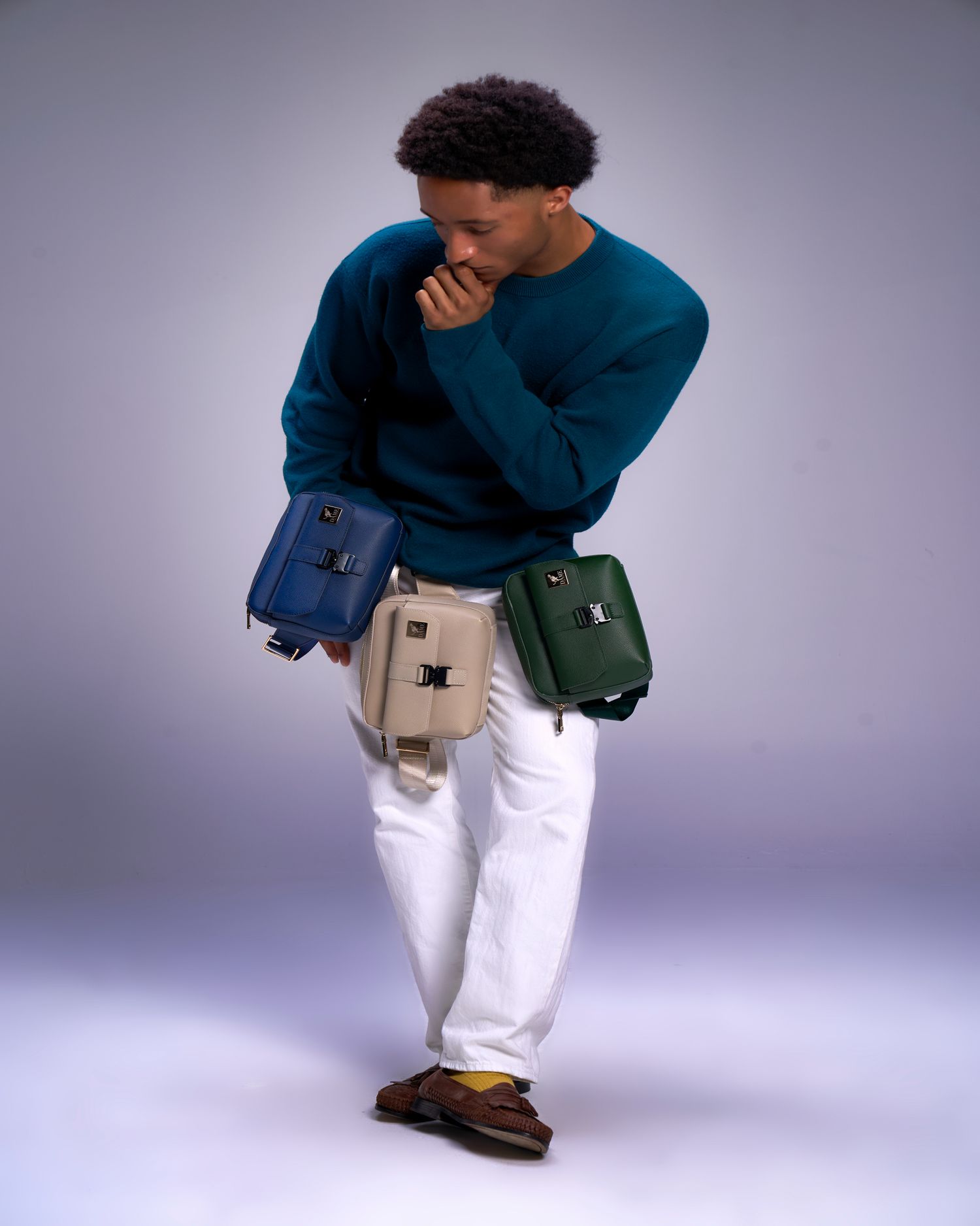 A person in a teal sweater and white pants models three different colored fanny packs against a gray studio backdrop.