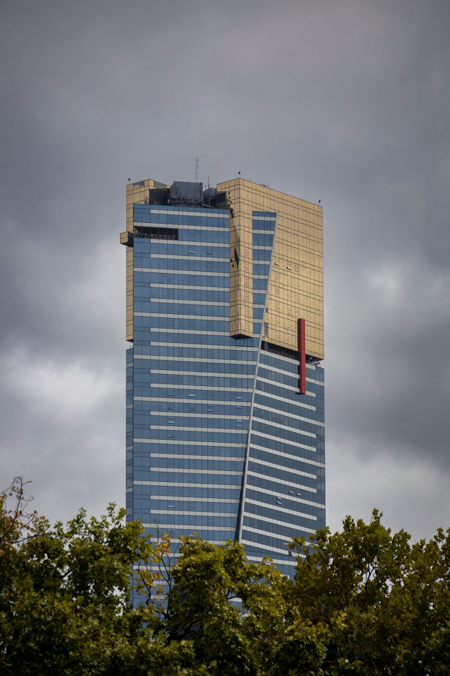 Tall blue-glass tower with gold accents piercing a cloudy skyline, standing bold above tree-lined streets.