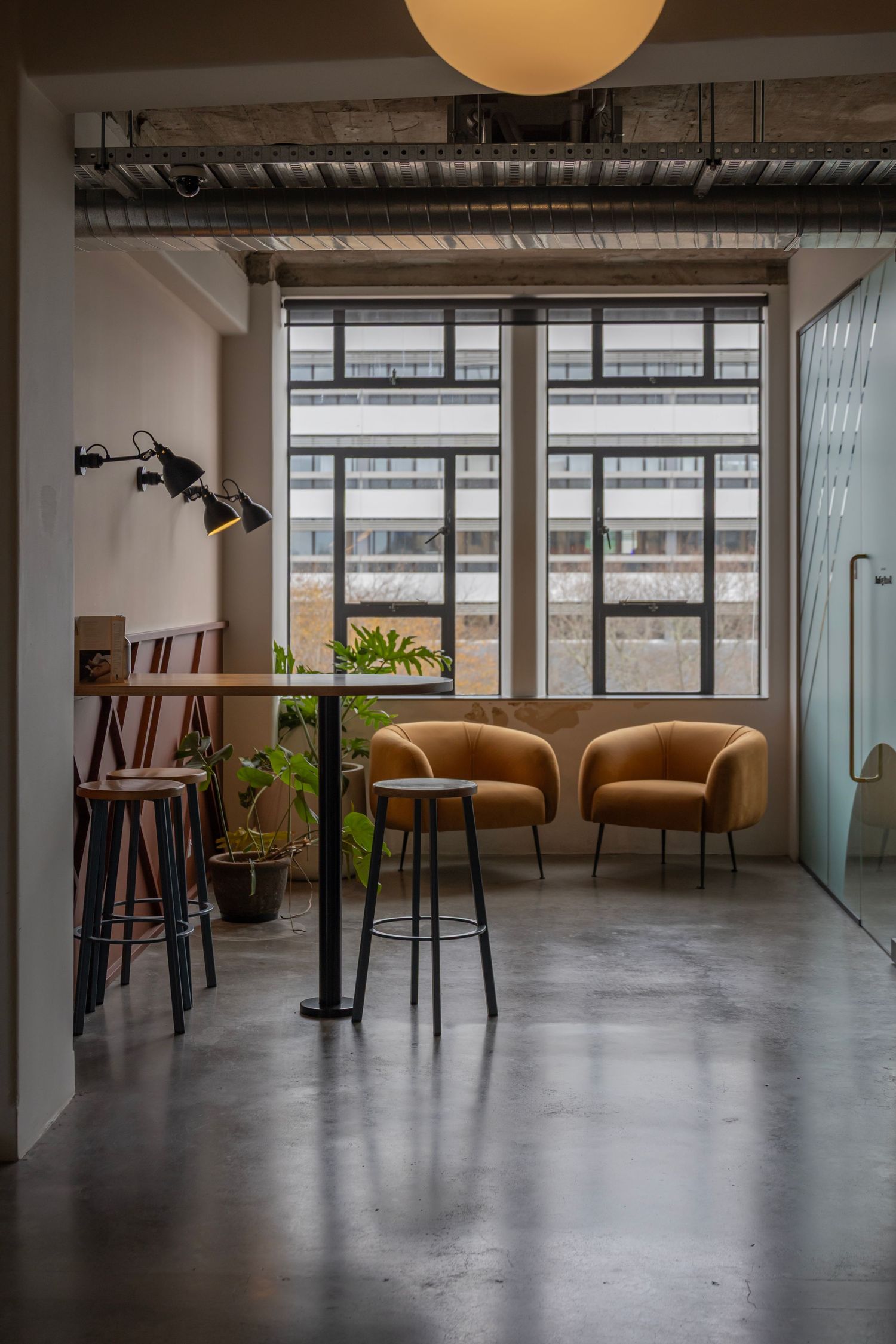 Small lounge with yellow chairs and bar stools under soft pendant lighting, framed by glass and concrete textures.