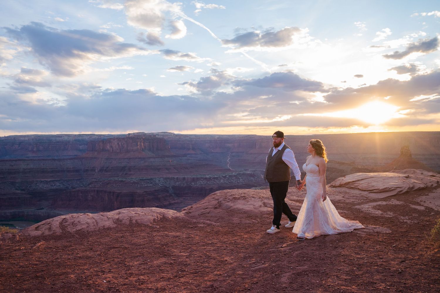 Bride and groom hold hands at canyon overlook during golden sunset in a breathtaking desert landscape.