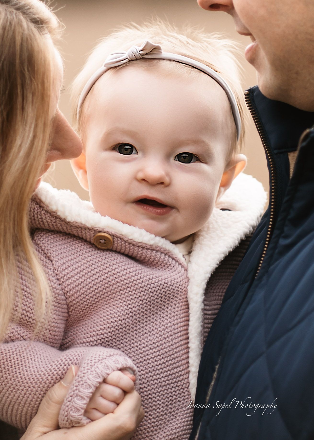 Baby wearing a cozy pink knit sweater smiles sweetly while being held in someone's arms.