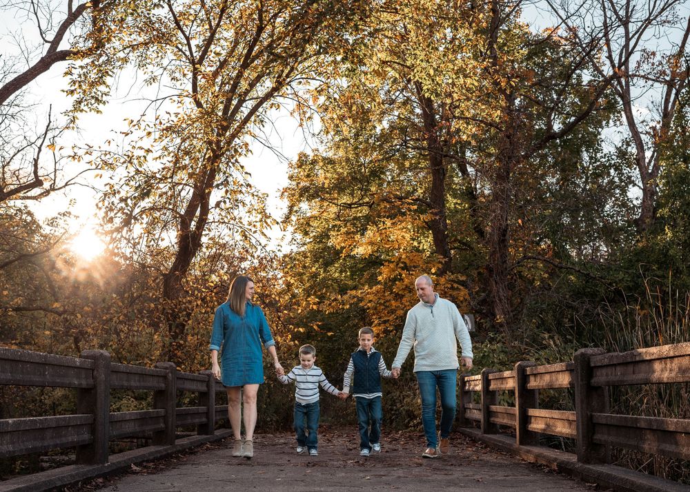 Family walking together on a wooden bridge through autumn trees at sunset in a park setting.
