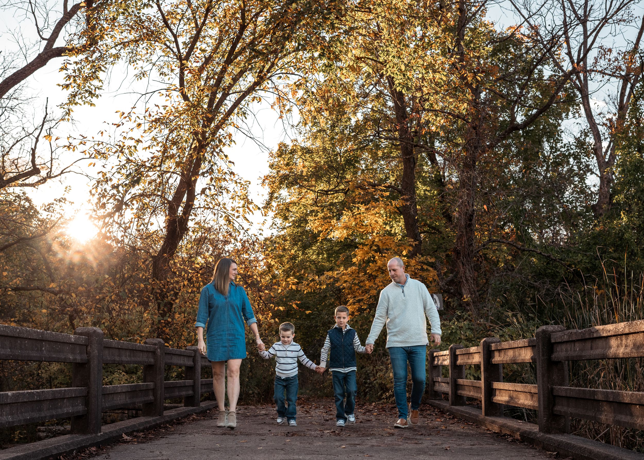 Family walking together on a wooden bridge through autumn trees at sunset in a park setting.