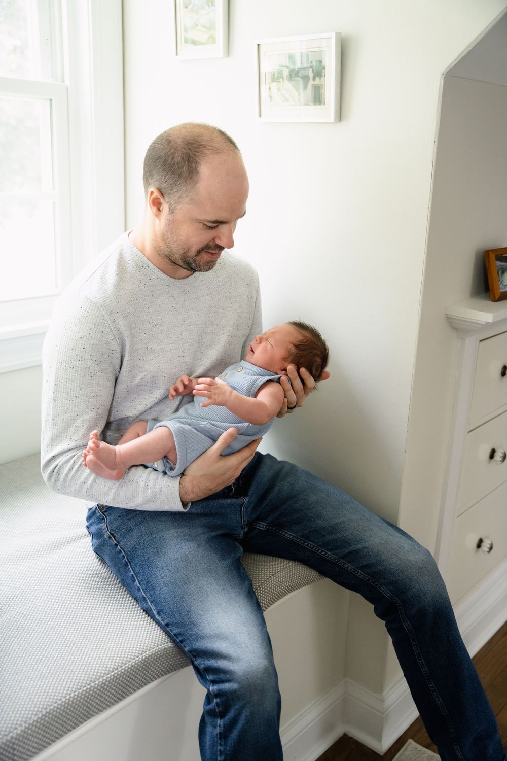 Parent in grey sweater and jeans sitting on window ledge holding and bonding with newborn baby.
