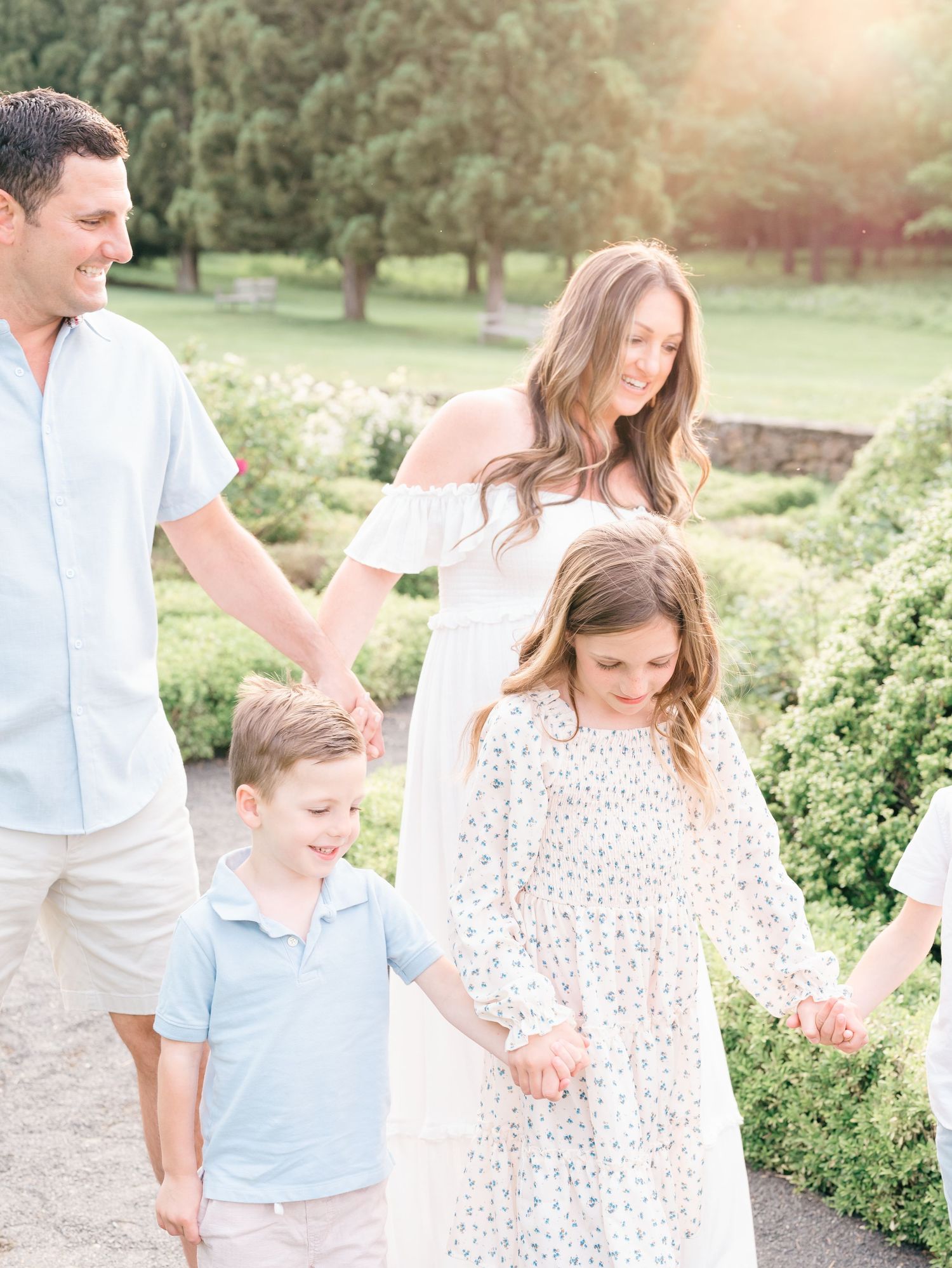 A family holds hands and walks through a garden in Middle town NJ with the sun rays touching them.