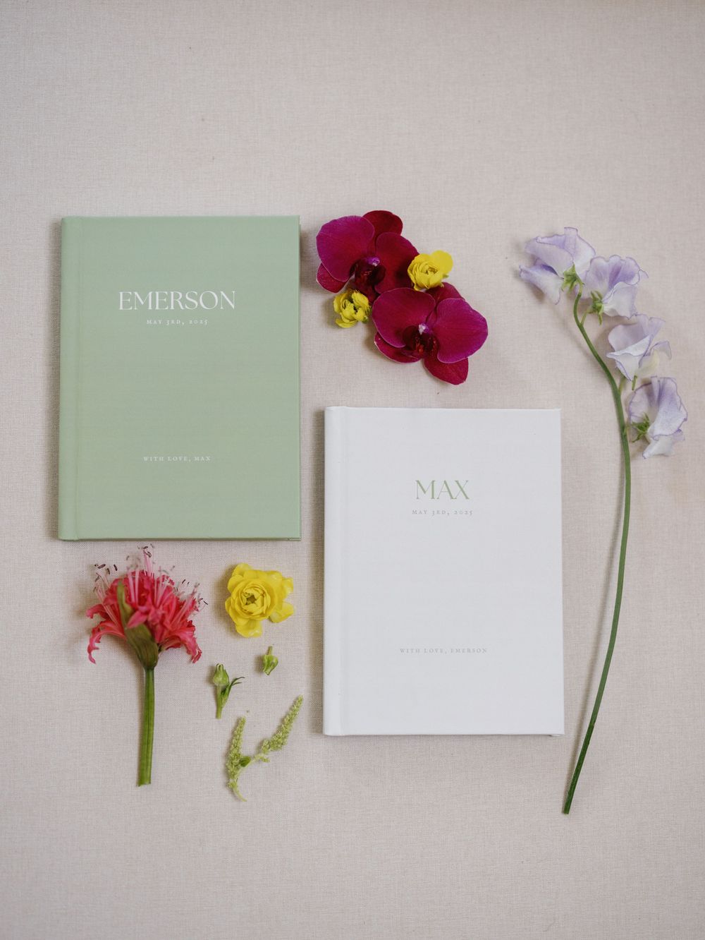 Wedding stationery flat lay with Emerson and Max books surrounded by colorful fresh flowers and orchids.