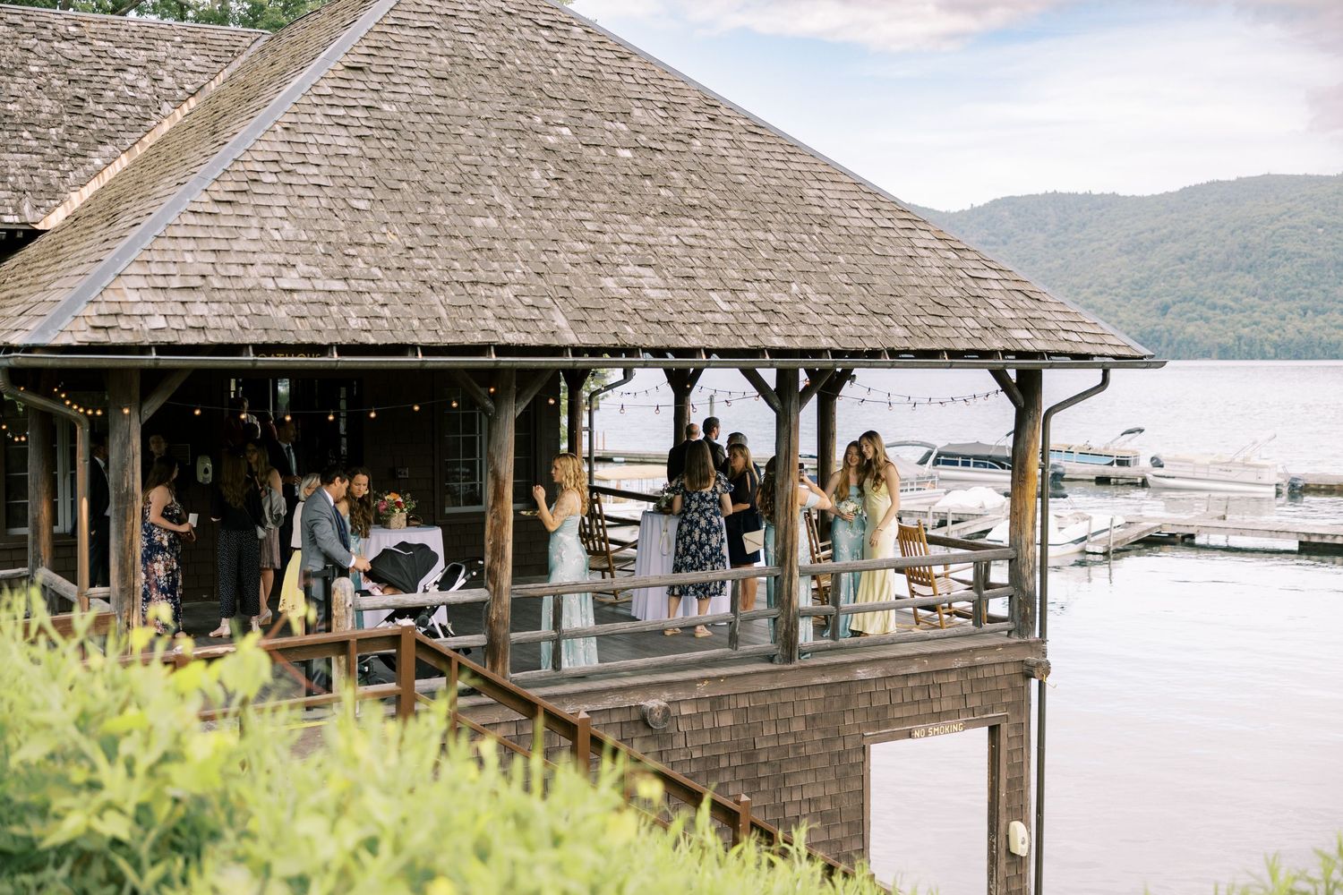 A rustic wooden boathouse overlooks Lake George with the Adirondack Mountains in the background on a cloudy day.