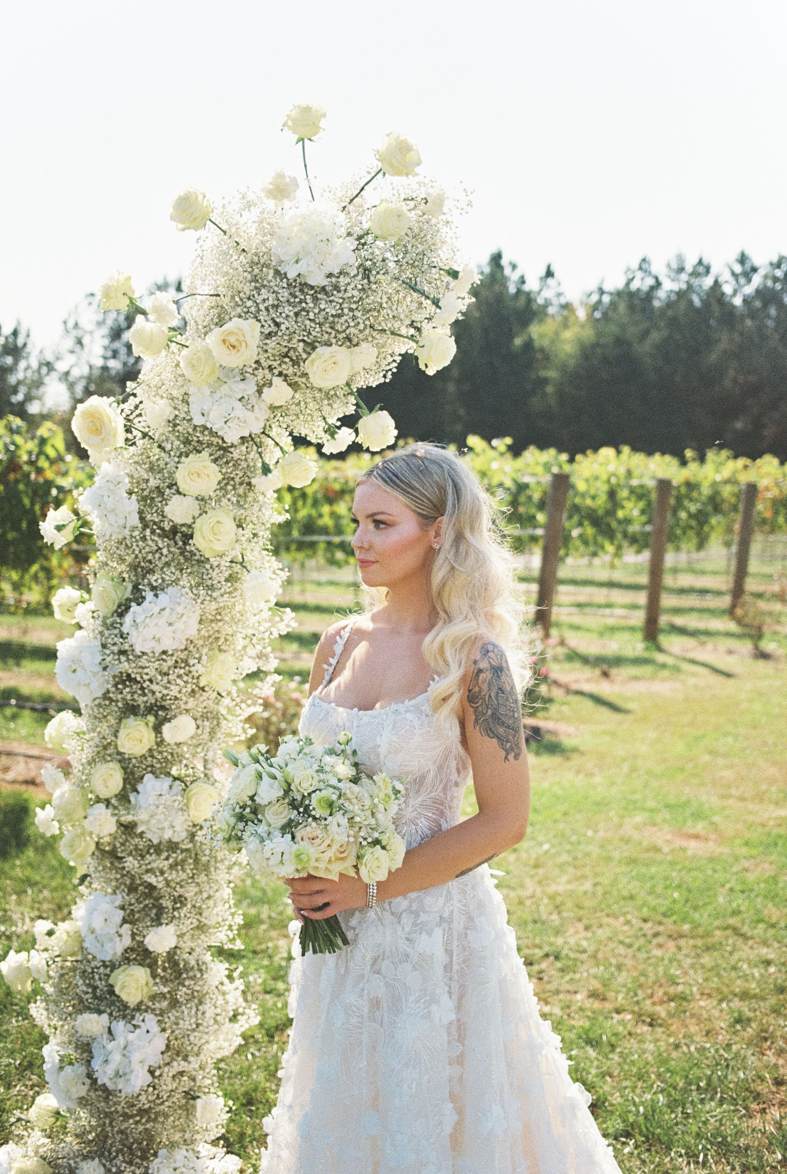 White floral wedding arch with roses and baby's breath in vineyard setting.