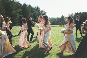 Wedding party dancing together on a sunny grassy field with pine trees in the background.
