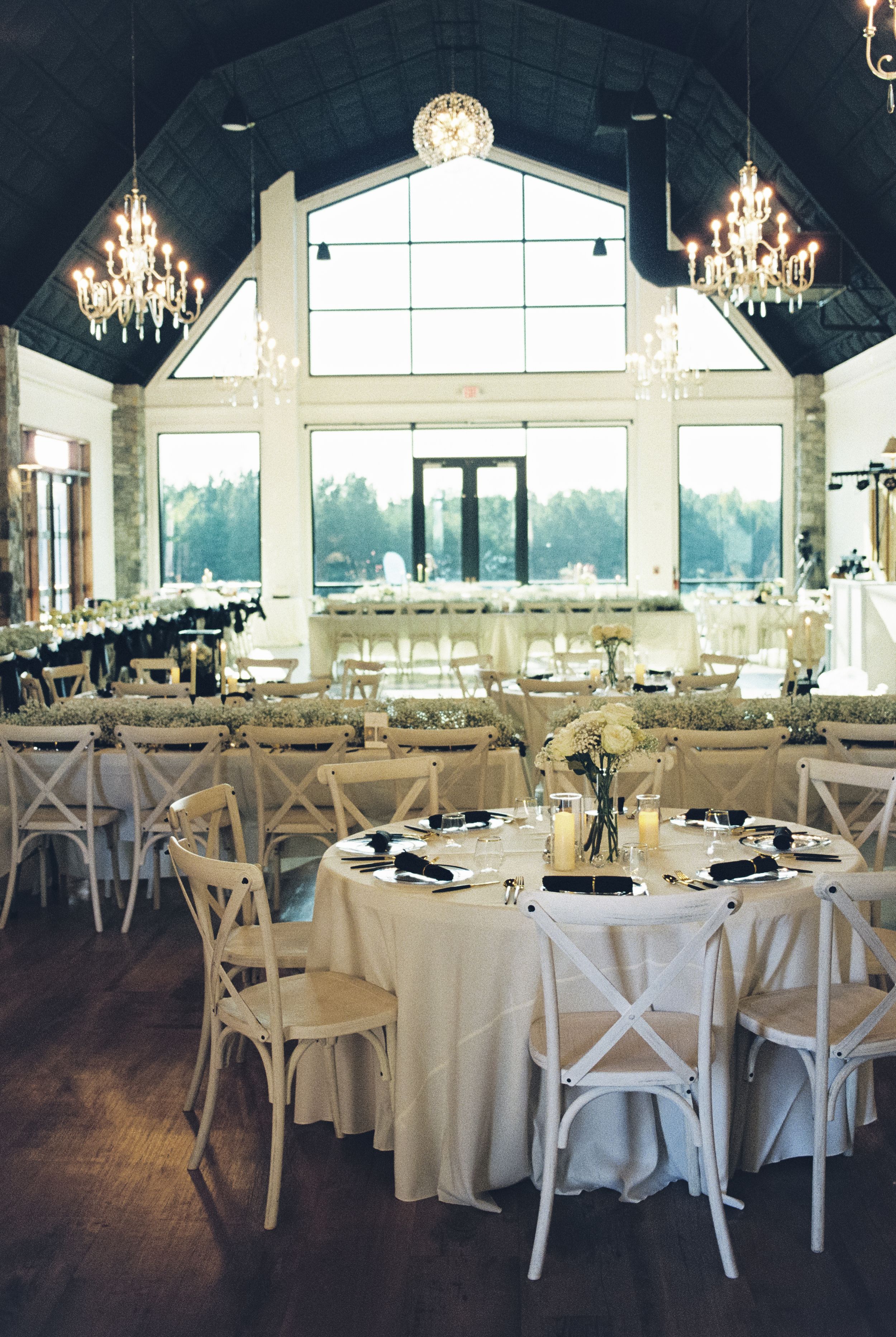 Elegant wedding reception venue with crystal chandeliers, white cross-back chairs, and floor-to-ceiling windows overlooking scenery.