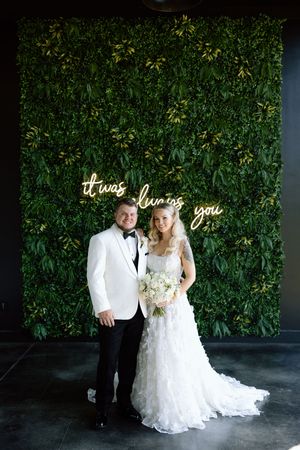 A couple poses in front of a lush green wall with illuminated 'it was forever you' neon sign at their wedding.