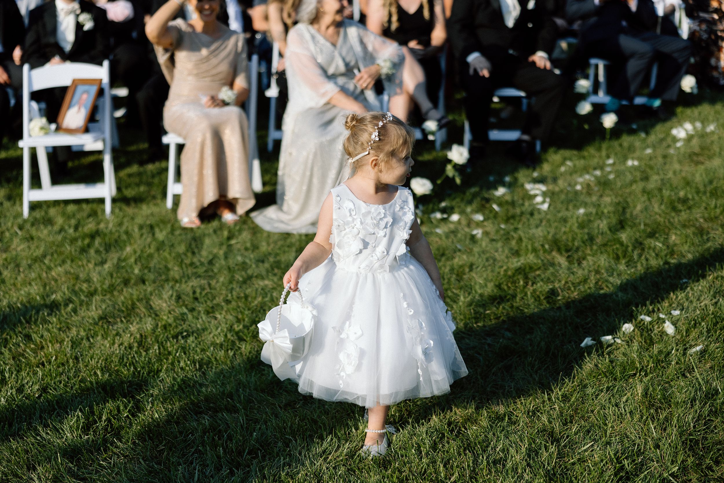 A young flower girl in a white tulle dress walks on grass during an outdoor wedding ceremony.