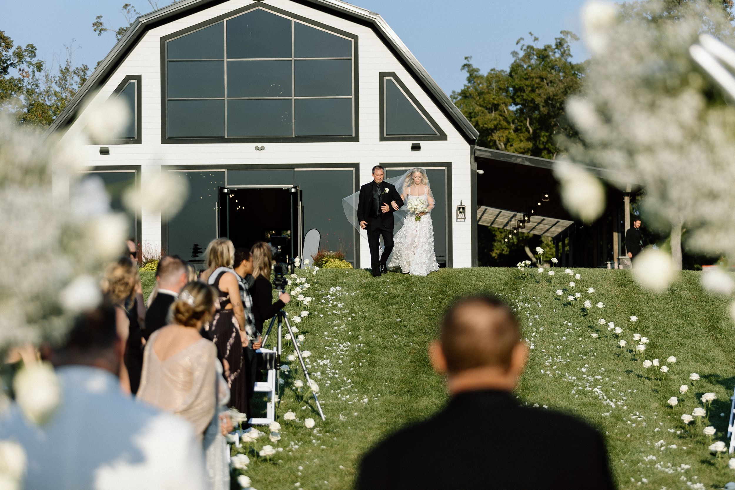 Wedding ceremony at a modern barn venue with white blossoms and guests seated on a grassy hillside.