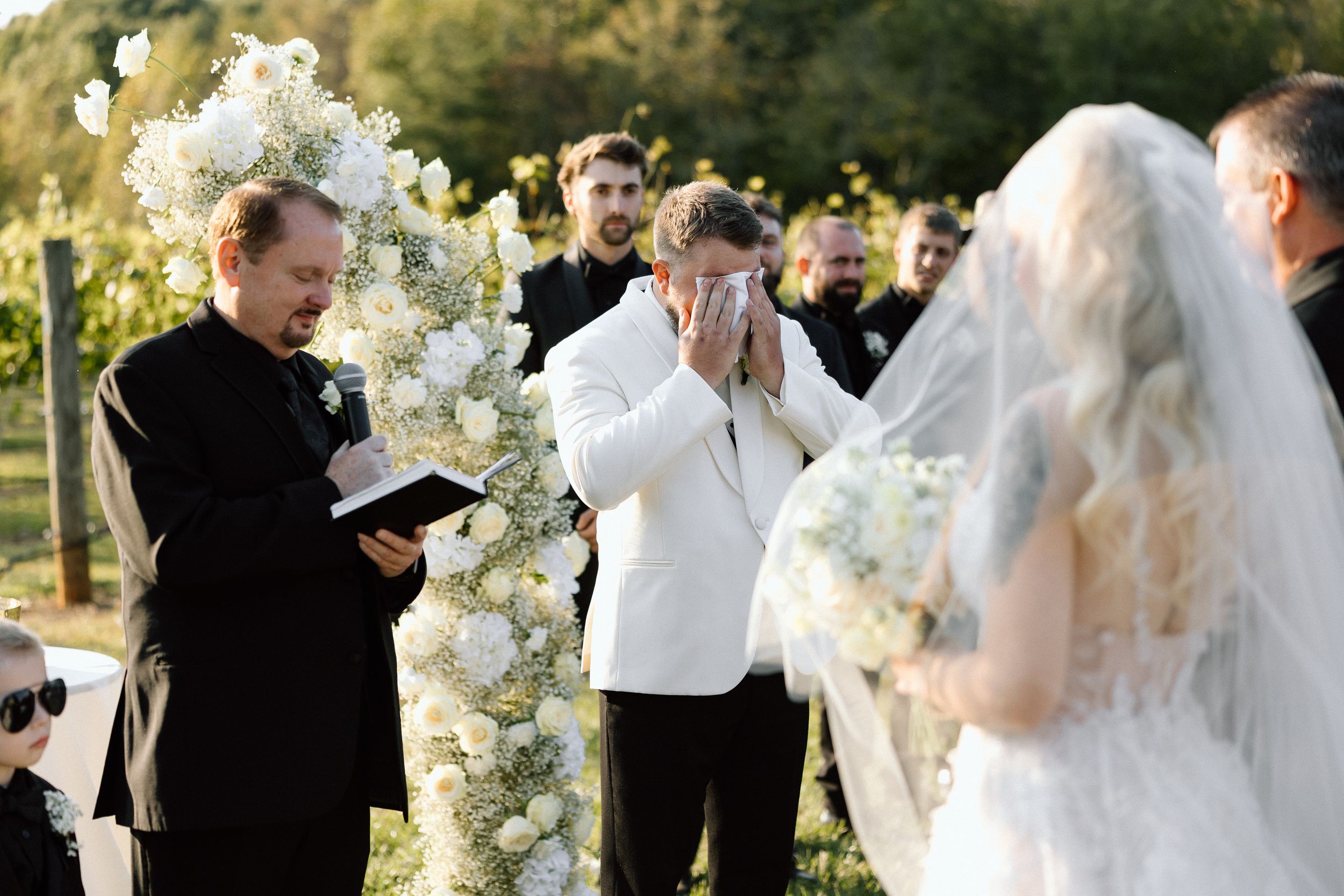 An emotional outdoor wedding ceremony moment with white floral decorations in the background.