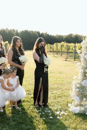 Wedding party standing by ornate white floral arch in a vineyard during golden hour.