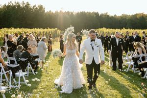 Newlyweds walk back down the aisle through cheering guests at a sunny outdoor ceremony with white chairs.
