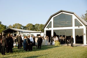 People gathered outside a white barn-style event venue with large glass windows for a reception on a sunny day.