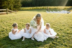 Beautiful scene of bride and flower girls in white dresses sitting together on sunlit green grass by countryside pond.