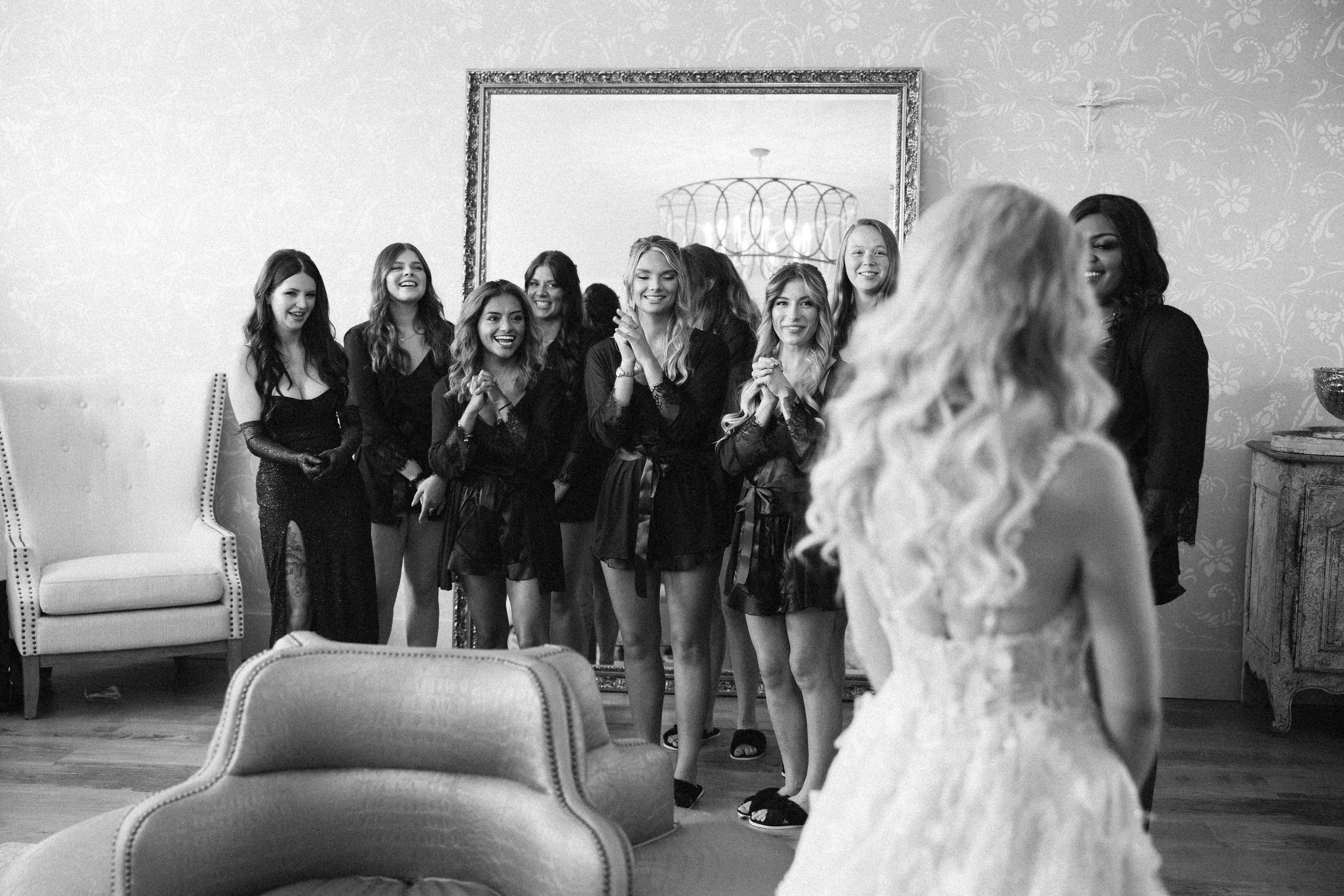 Black and white photo of wedding party in matching black robes gathered in elegant room.