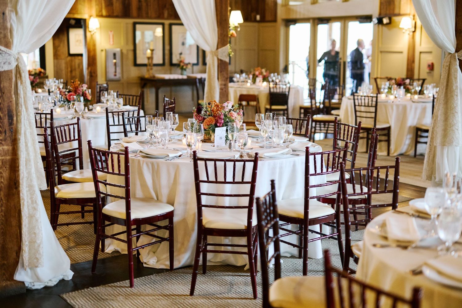 Elegant wedding reception venue with dark wooden chiavari chairs and round tables set with white linens and floral centerpieces.