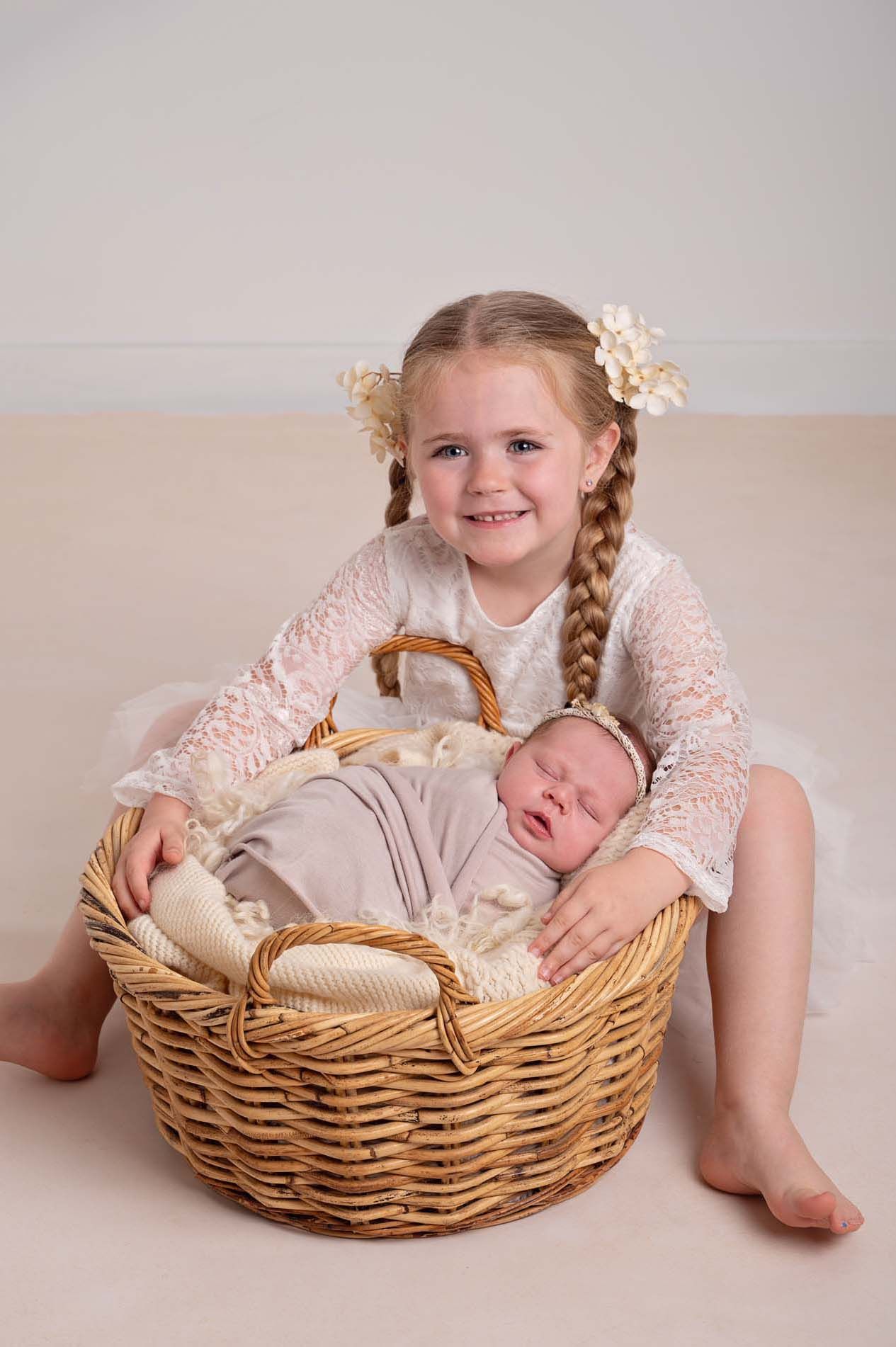 Five-year-old girl smiles sitting holding a natural basket prop with her sister sleeping in it during a sibling newborn photo session.