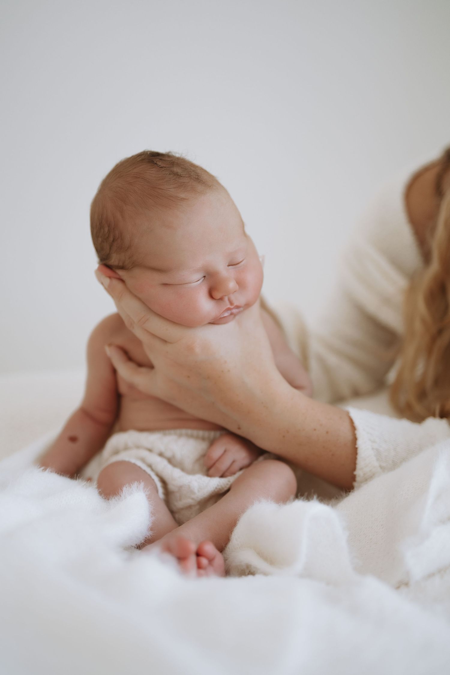 Curled up newborn resting on cozy white bedding in gentle natural light.