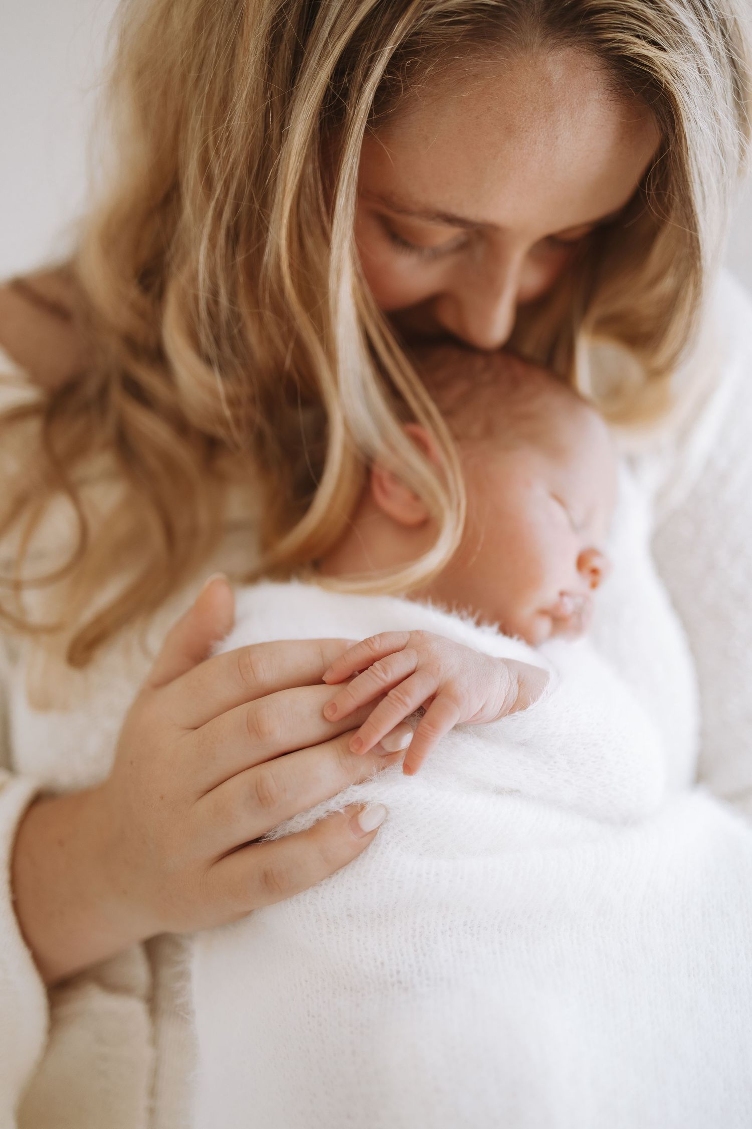 Tender moment captured in soft lighting showing emotional parent dressed in white holding sleeping infant.