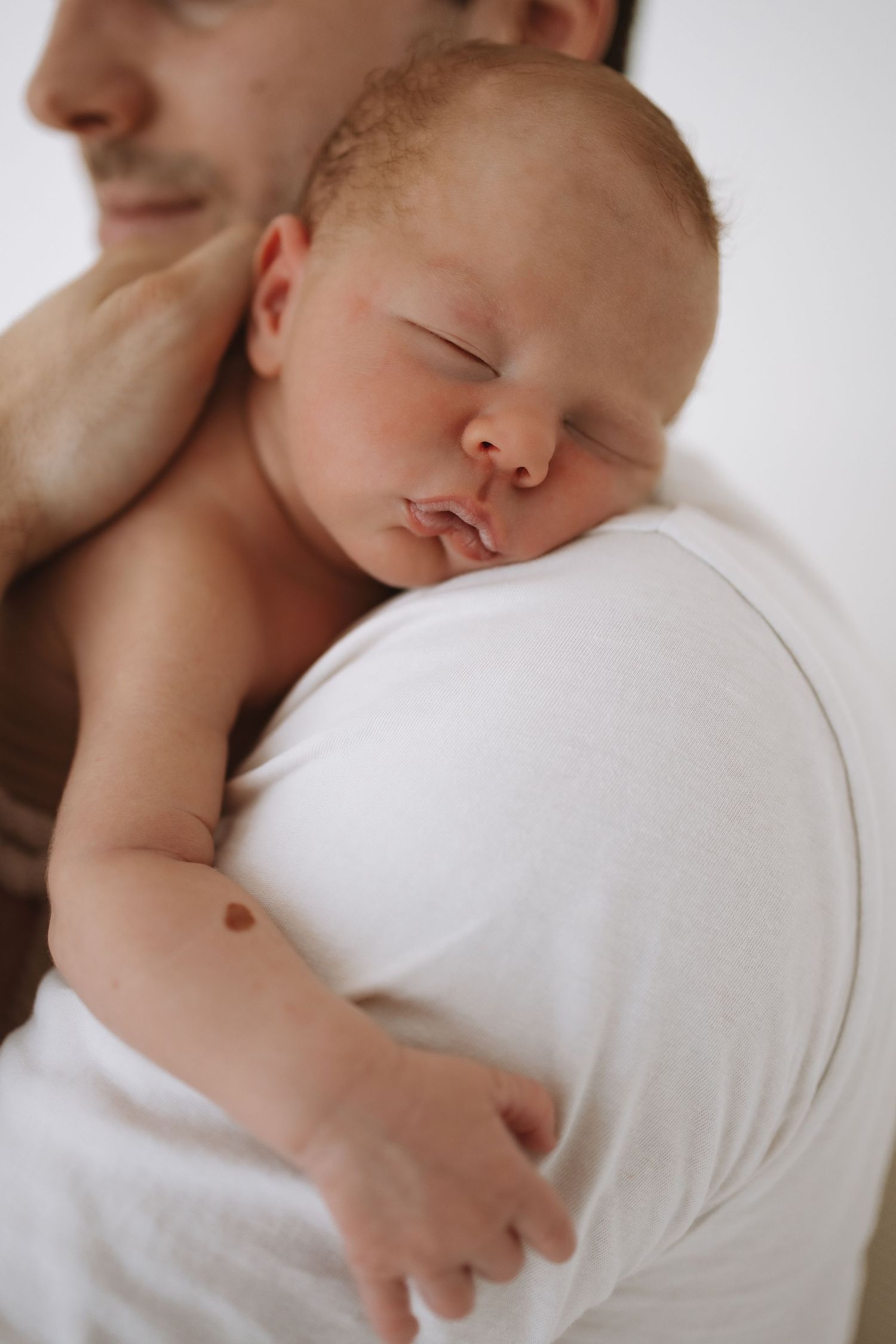 Newborn baby sleeping peacefully on parent's shoulder in white outfit against light background.