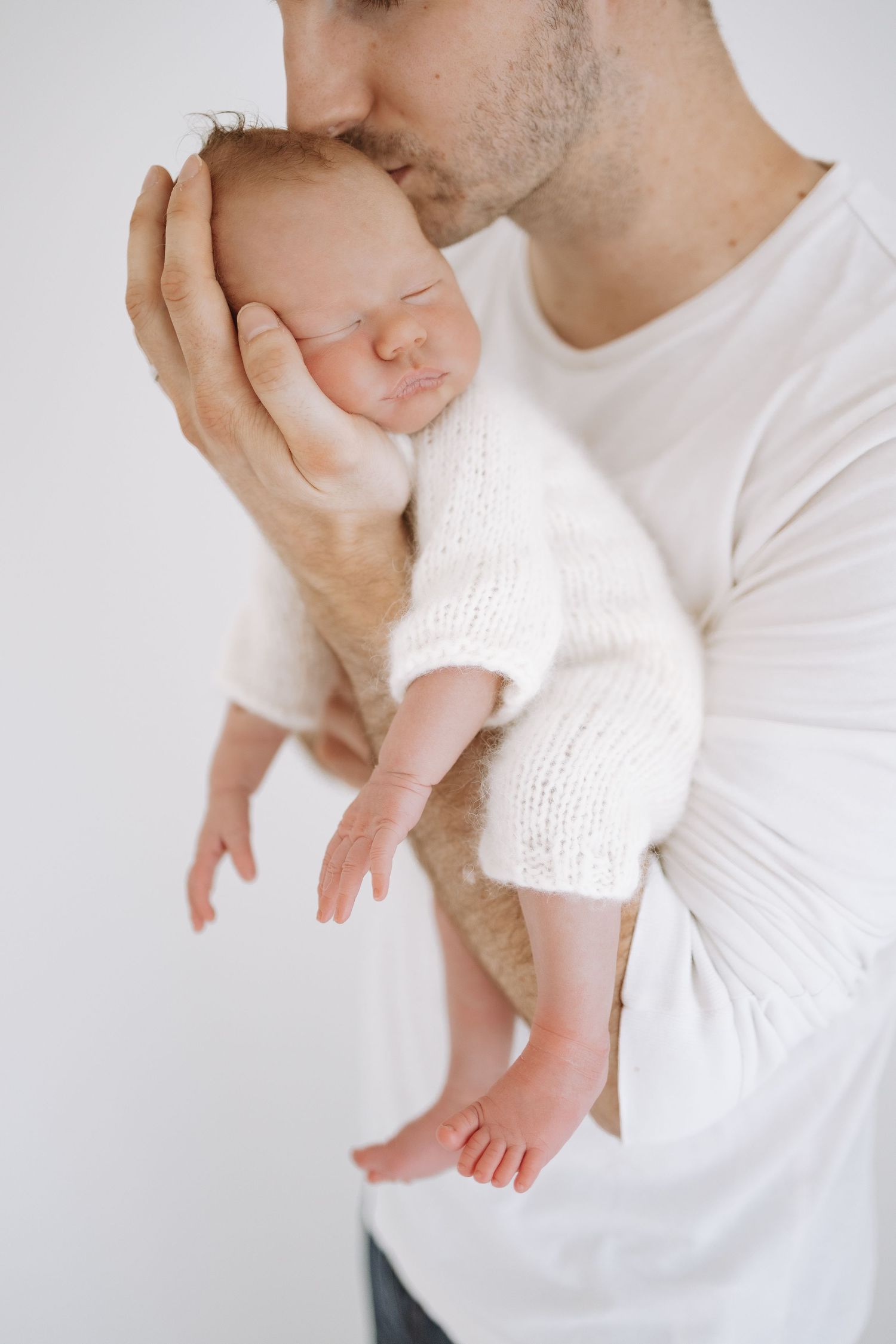 Tender moment showing newborn infant in white knitted sweater being cradled gently against white background.