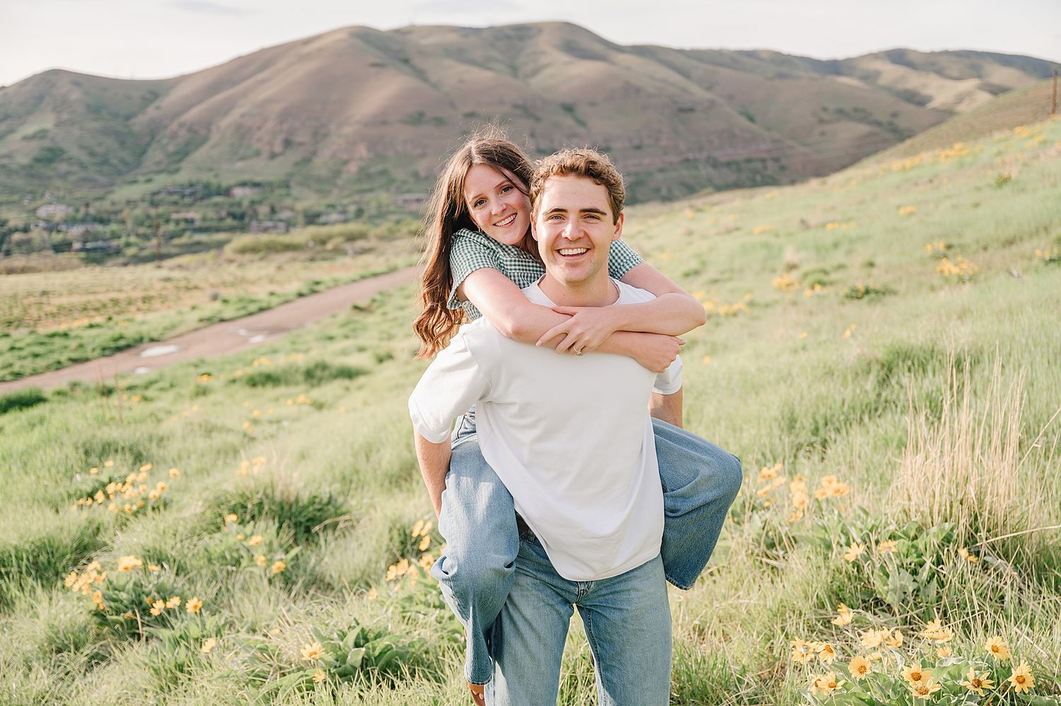 Happy couple sharing a playful piggyback moment against scenic mountain backdrop with lush green grass.