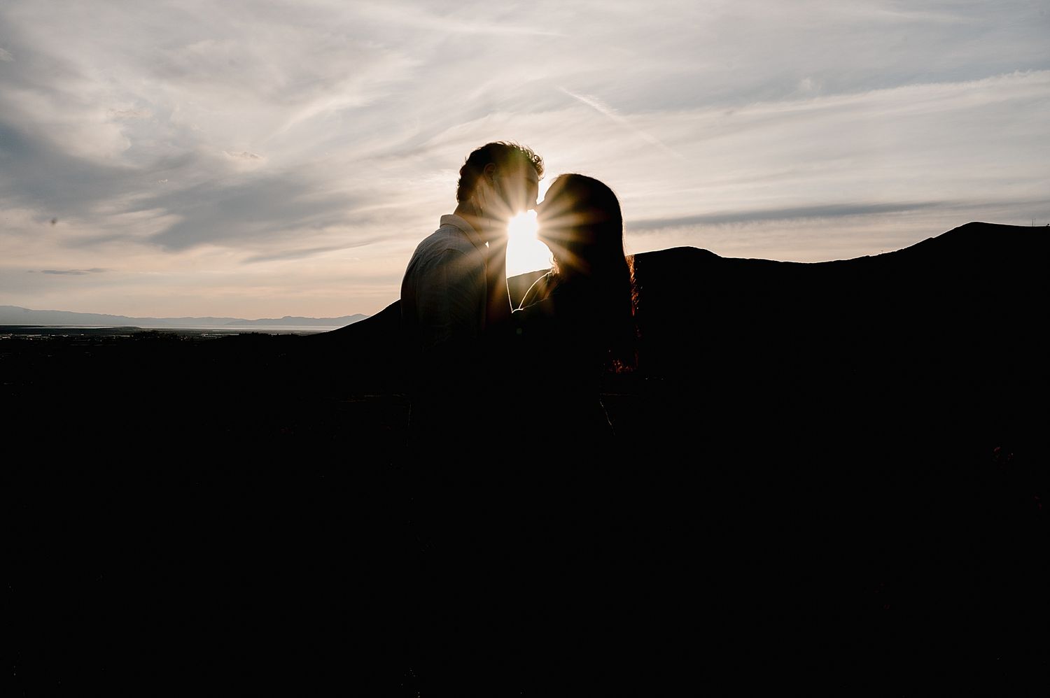 A silhouetted couple shares an intimate moment as the sun sets behind mountains creating a dramatic backlit effect.