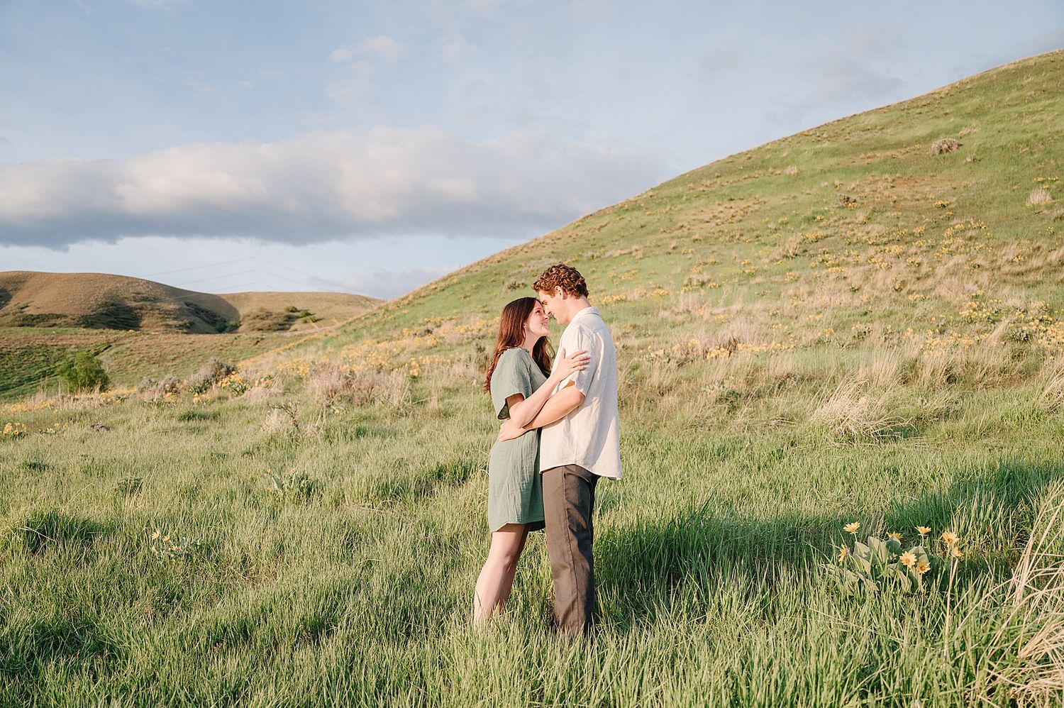 A couple embraces in a serene meadow against rolling green hills and cloudy skies at golden hour.