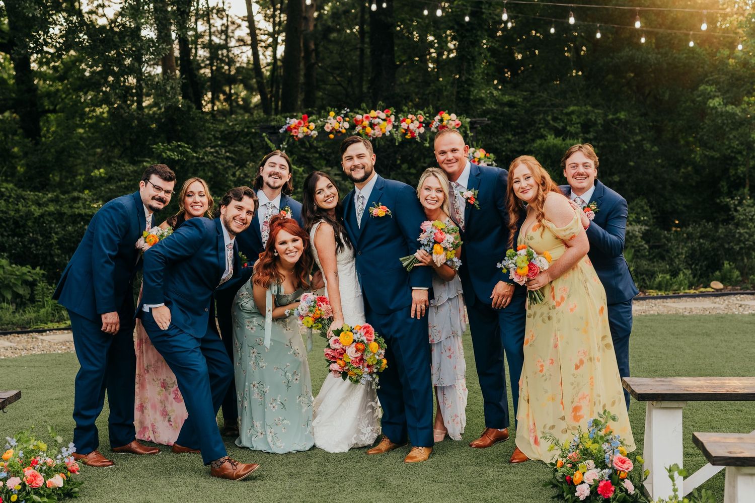 Wedding party stands together outdoors at sunset wearing navy blue and pastel attire with autumn-colored floral bouquets in Longview, Tx.
