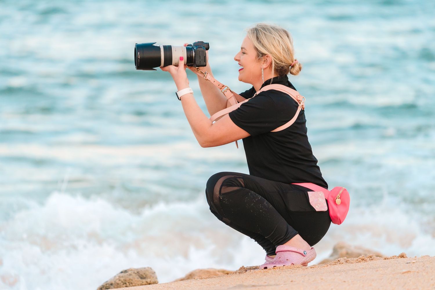Photographer in black attire taking photos on beach with professional camera.