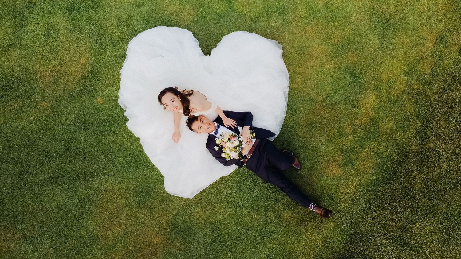 Aerial drone shot of bride and groom lying on grass forming heart shape with wedding dress.