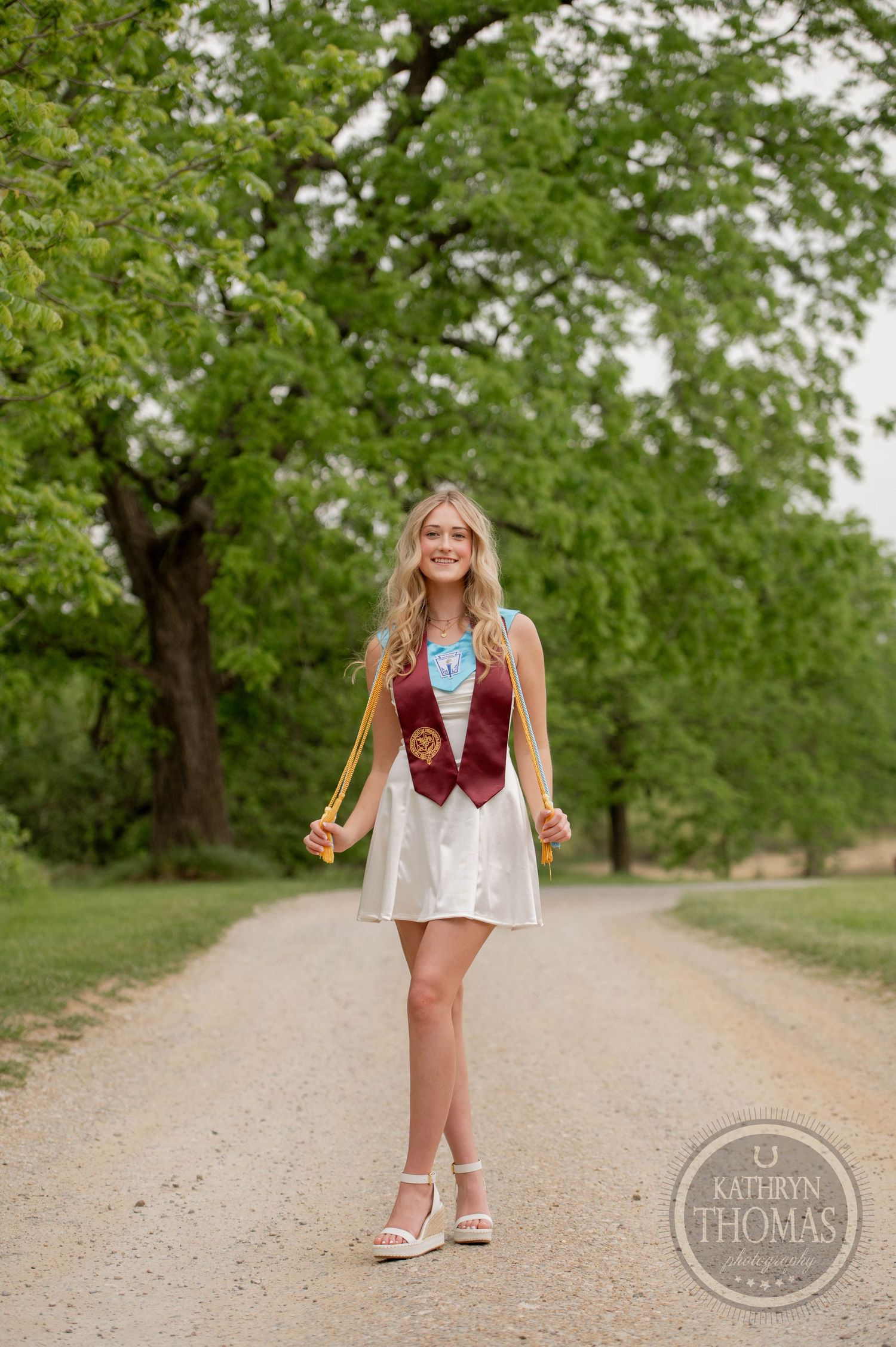 A fashionable outfit featuring a short white dress, burgundy vest and sandals on a peaceful dirt path surrounded by green trees.