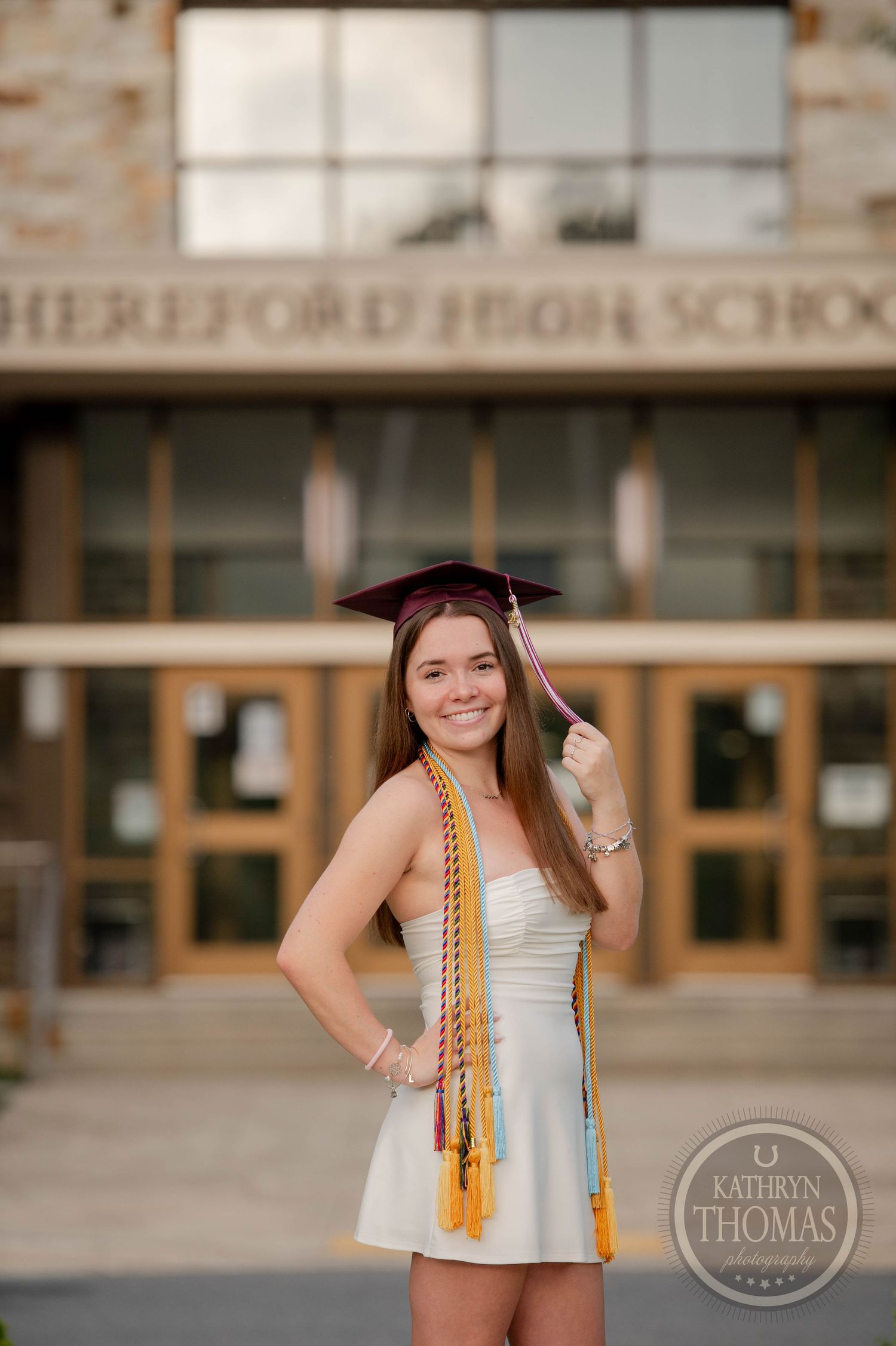 A student wearing a white dress and graduation cap poses in front of their high school building.